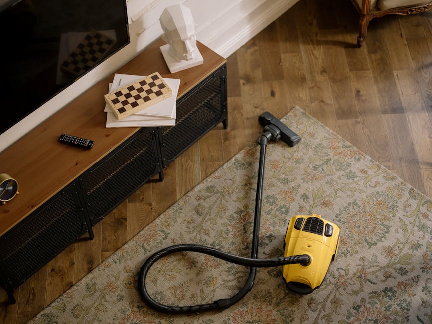 A yellow canister vacuum cleaner with a black hose is placed on a decorative carpet in a living room, indicating a surface cleaning process. The room features wooden parquet flooring, a black media co