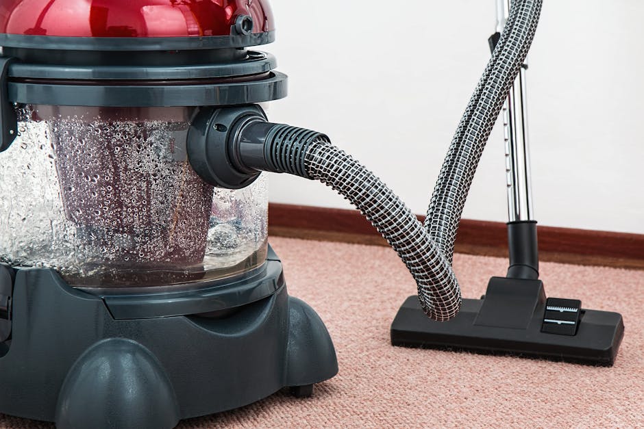 A yellow canister vacuum cleaner with a black hose is placed on a decorative carpet in a living room, indicating a surface cleaning process. The room features wooden parquet flooring, a black media co