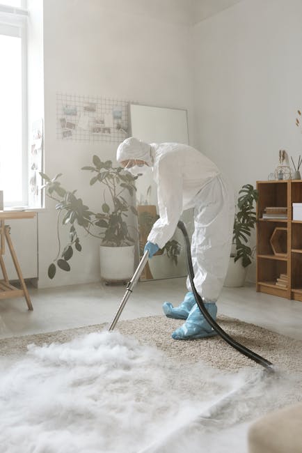 A person dressed in full protective cleaning gear, including a white suit, gloves, face mask, and blue shoe covers, is performing deep cleaning on a beige carpet in a well-lit, modern living room. The